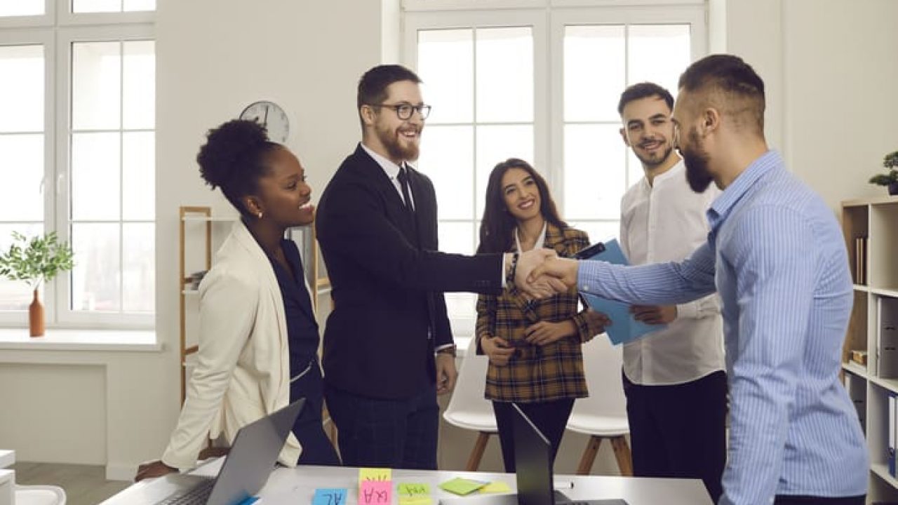 Male leader of a multiracial business team greets a new team member shaking his hand.