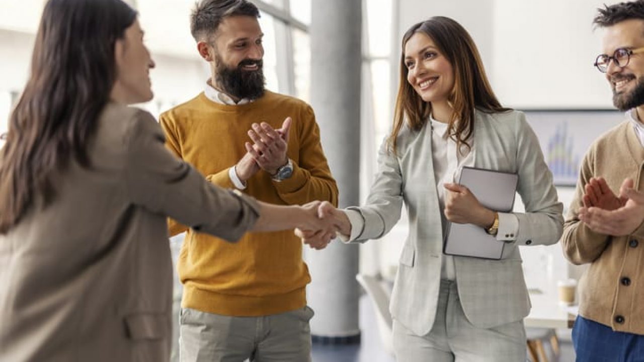 Two successful businesswomen shaking hands and closing deal and partnership while businessmen applauding them.