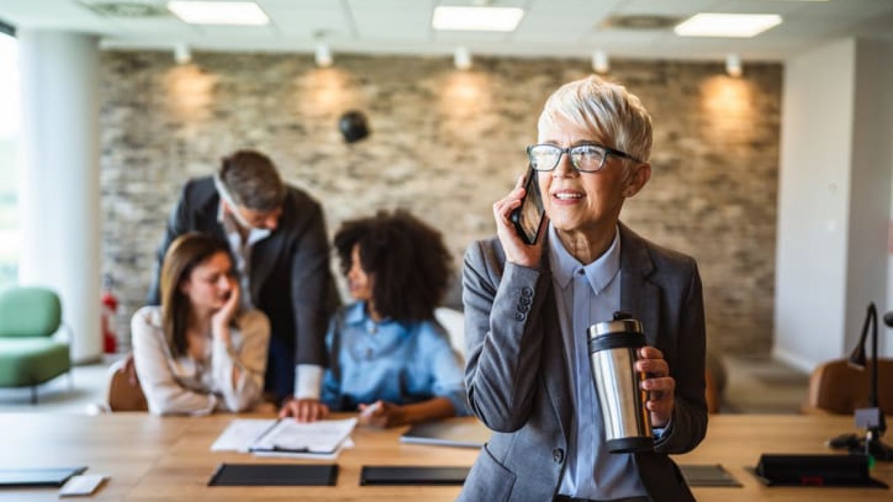 aged businesswoman talk on mobile phone and drink coffee at workplace