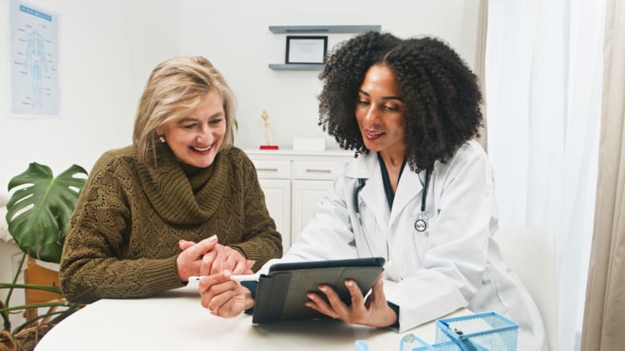 Friendly Doctor Consults Older Woman With Tablet In Clinic, Careful Health Advice And Wellness Support